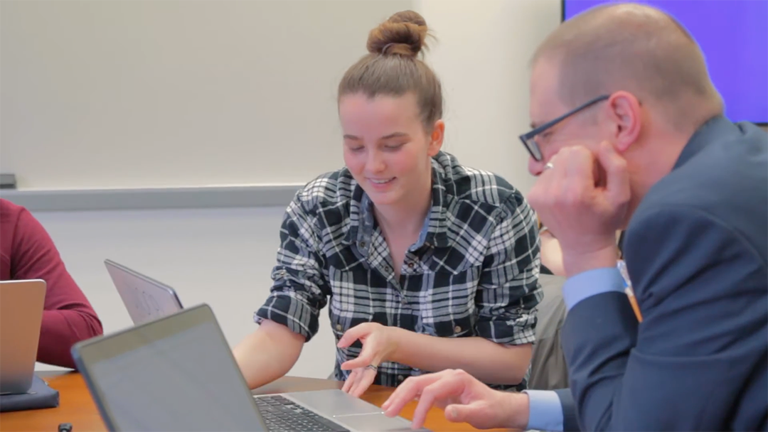 Student and faculty member working together at a table with laptops. The student is smiling and gesturing toward the screen while the faculty member listens.'