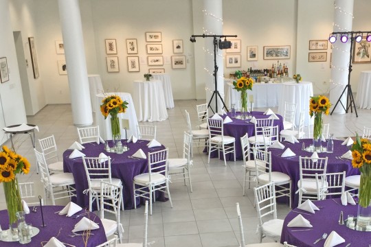 The university gallery space set up with purple table-clothed tables with sunflower bouqets.