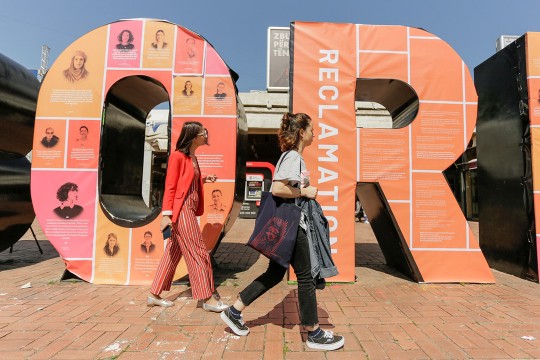 Two people walk by sculpture of letters O and R covered with images of Kosovar women.