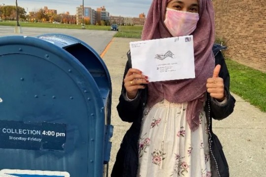 a voter holding her mail-in ballot next to a USPS mailbox.