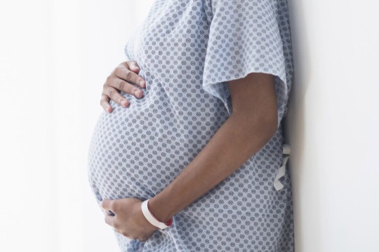a pregnant person wearing a hospital gown standing against a wall.