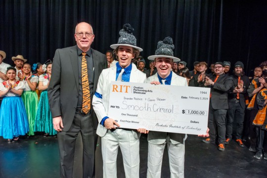 David Munson, Jr., RIT President, stands on stage with 2 students that won the $1,000 prize. Other performers stand behind them