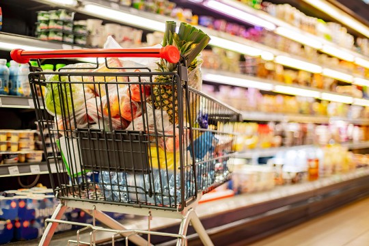 A grocery cart filled with perishable foods like fruits and vegetables standing alone in the dairy aisle of a grocery store. The background of the aisle has a blurred effect.