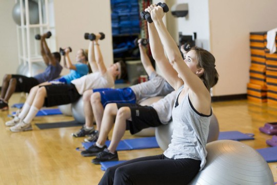 students workout in a gymnasium setting holding weights overhead.