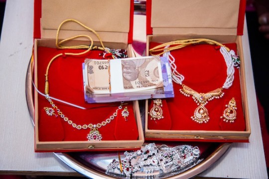 Two open jewelry boxes display gold necklaces and matching earrings on red velvet, with a stack of currency placed between them.
