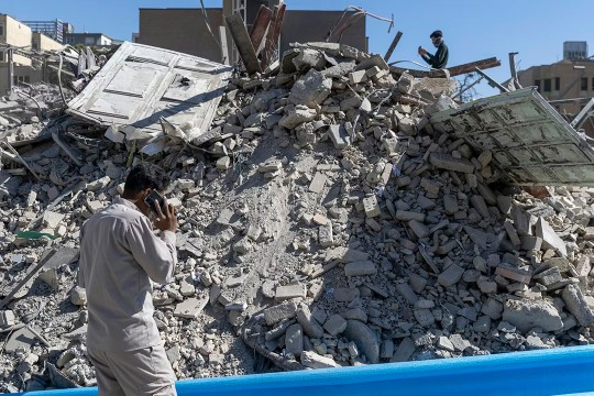 Rubble from a police station damaged in airstrikes on March 3, 2026 in Tehran, Iran. A man in the foreground is seen with his back turned talking on the phone. Another man stands on the top of the pile of rubble taking photos with his phone.