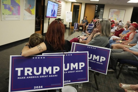 people sit in a large room holding Trump Twenty Twenty Four signs on their laps.
