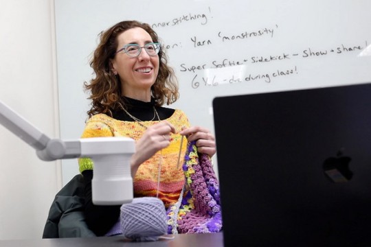 a woman with dark curly hair and glasses crochets at the front of a class in front of a whiteboard