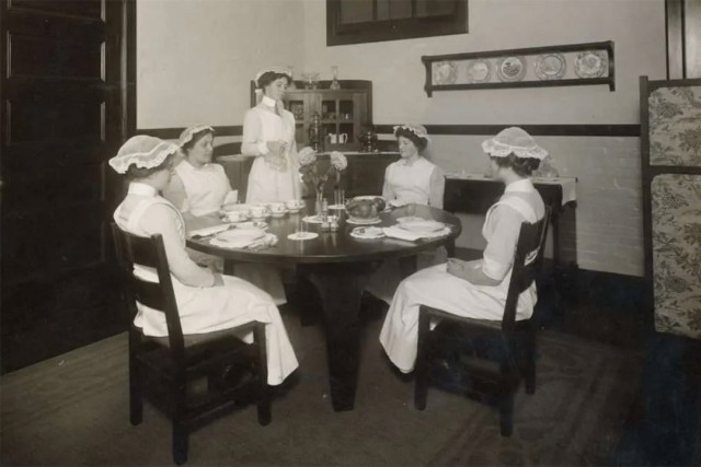 a historical photo of four women seated and one standing around a dining room table.