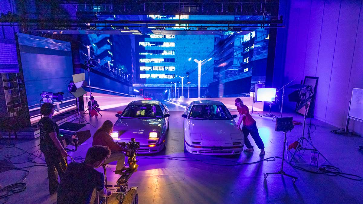 a film production set with two cars on the floor and a road scene on a screen in the background.