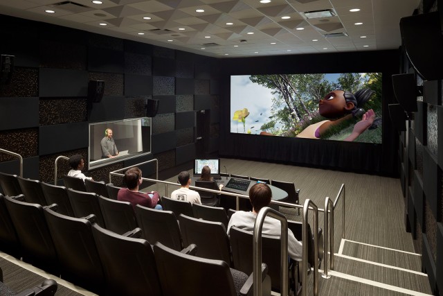 Student sits at table with controls while another student sits at electric keyboard and screen in background with controls projected onto it