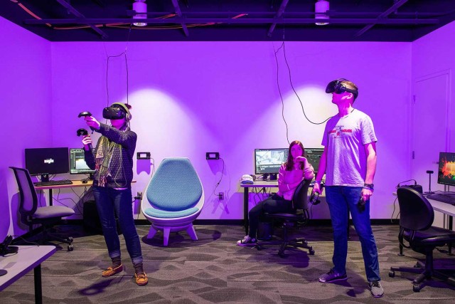 Two students stand wearing VR goggles and holding controllers in room with chairs and computers