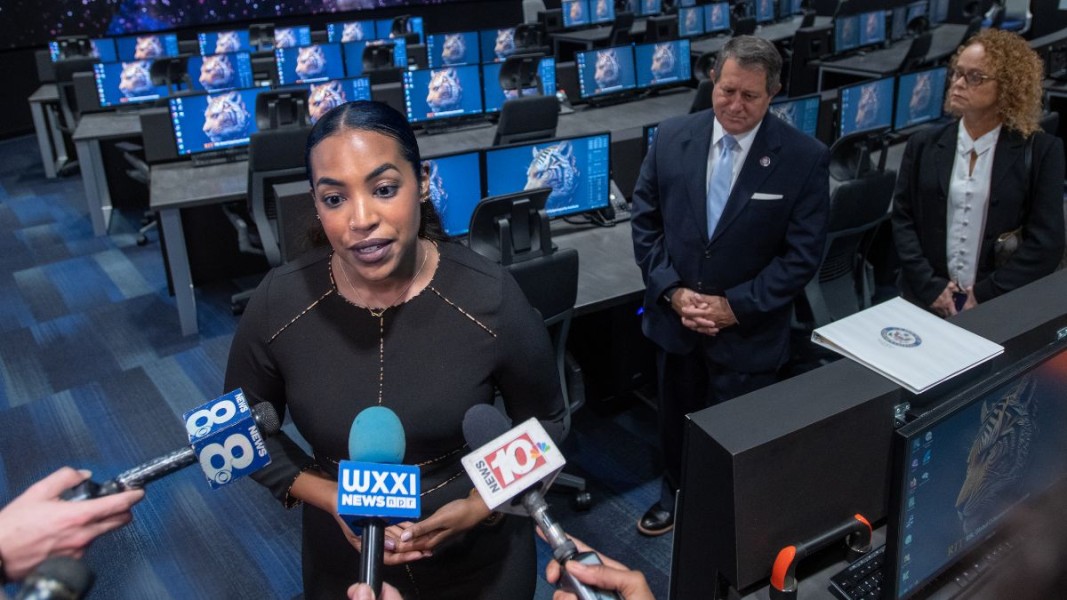 A press conference in a high-tech control room features a speaker addressing reporters and news microphones.