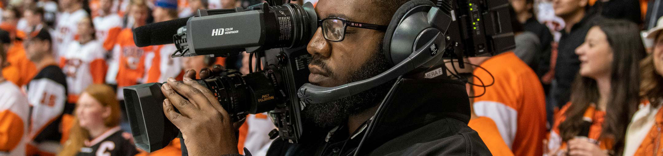 Camera operator holding a camera on his shoulder with RIT fans in orange in the background