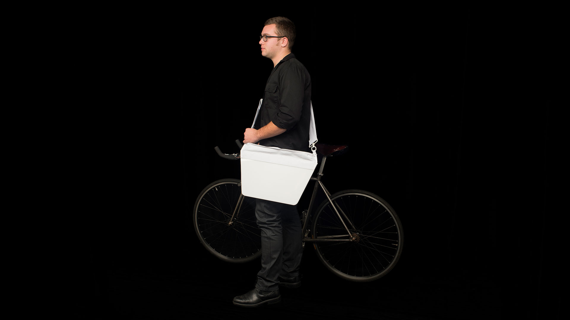 Student standing facing left with bike on his right side, a white messenger bag across his body with a tapered flap design