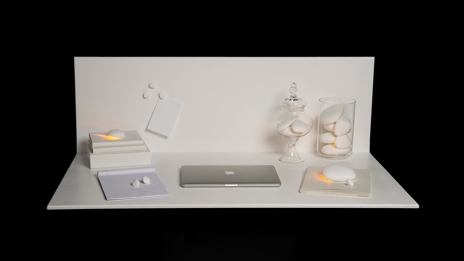 white desk surface with white back wall, laptop and assorted stacked papers on desk, white task stones of various sizes on different papers, some in clear glass containers, three small task stones on back wall, some task stones shining light from bottom