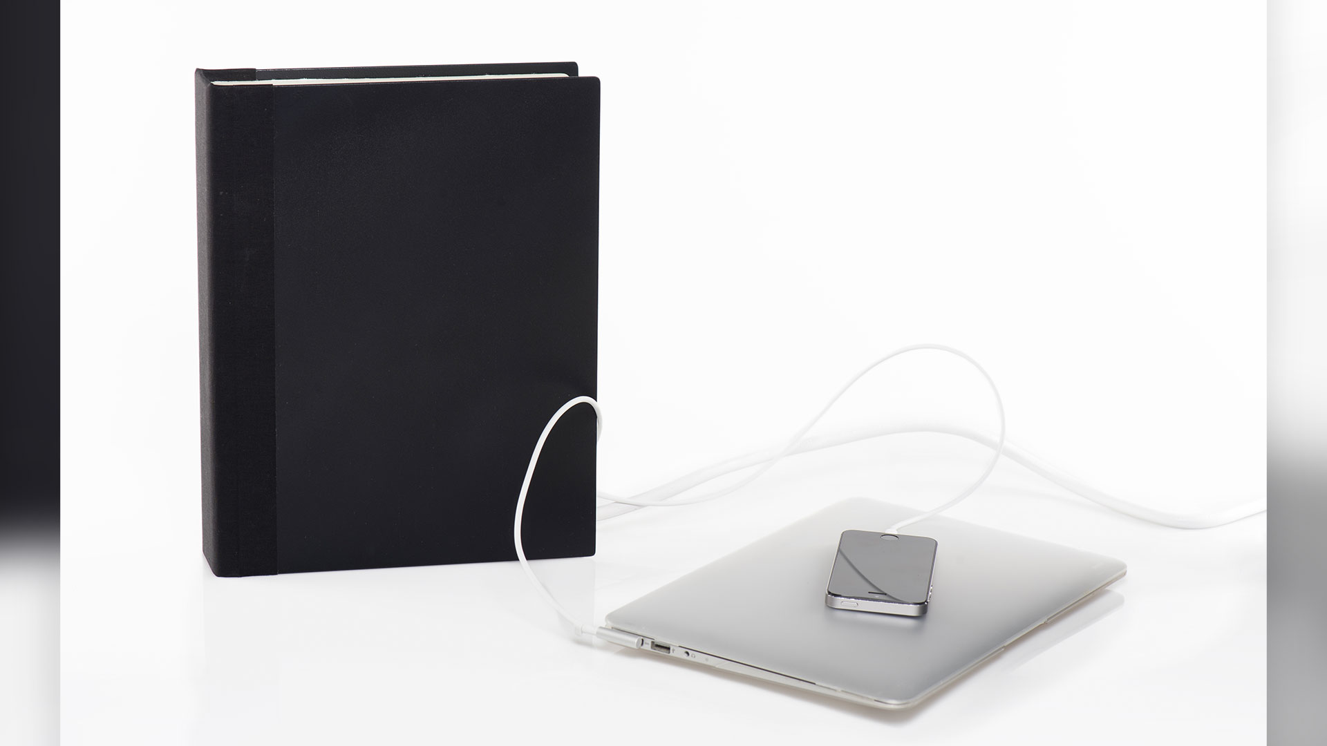 Black hardcover book standing to the left of laptop and phone both plugged into cords that extend into side of book