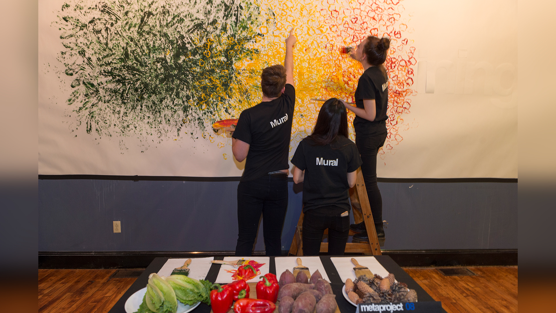 Three students standing in front of long white wall stamping paint on with various food items