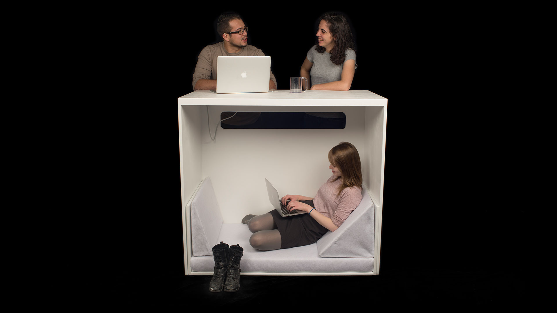 Woman inside desk space of Personal Space Bar with bench component inverted for a lounging area, woman works with laptop inside while above at desk space on top of compartment two students stand talking to each other with a laptop out 