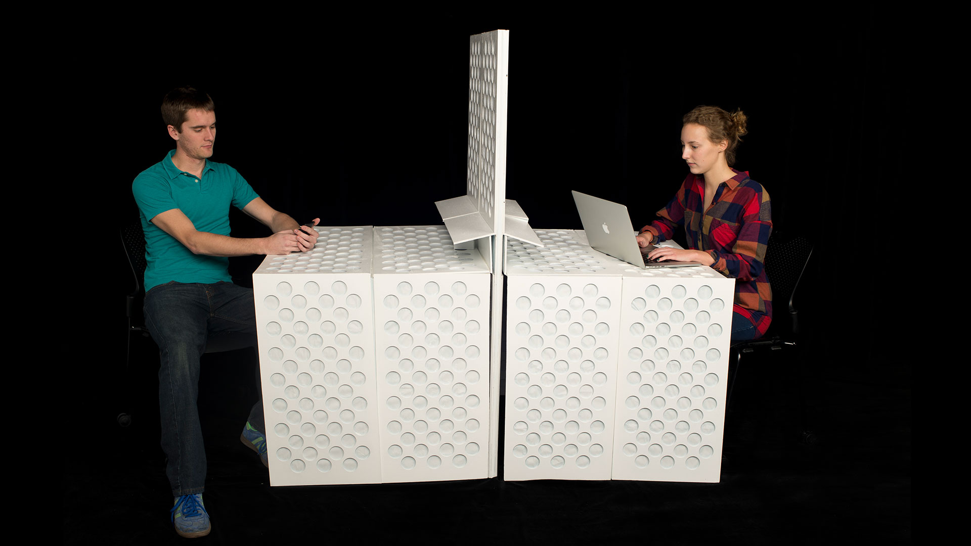Two students on either side of room divider, each with a section folded down for their own mobile desk, one student on phone and one student on laptop 