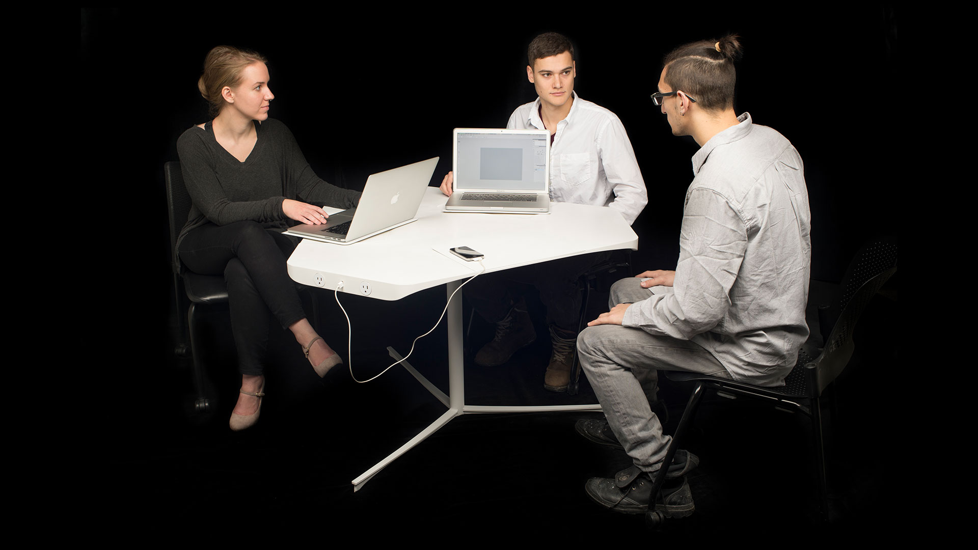 Three students around table, triangularly geometric white table with one flat side, two students on other sides with laptops plugged into outlets attached to table