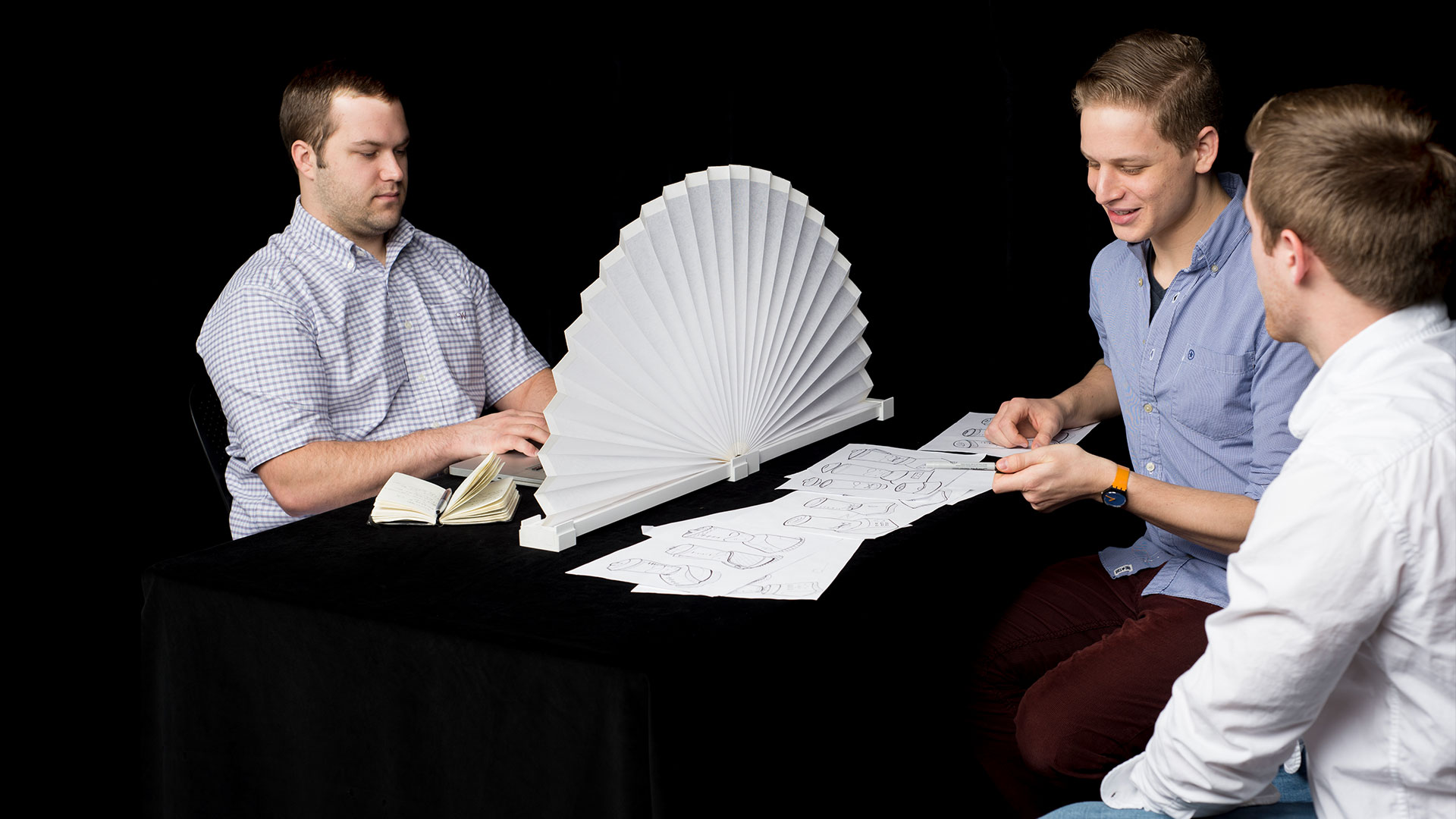 Small white table fanned open with accordion pleating connected to white plastic base, students on either side of divider doing separate tasks