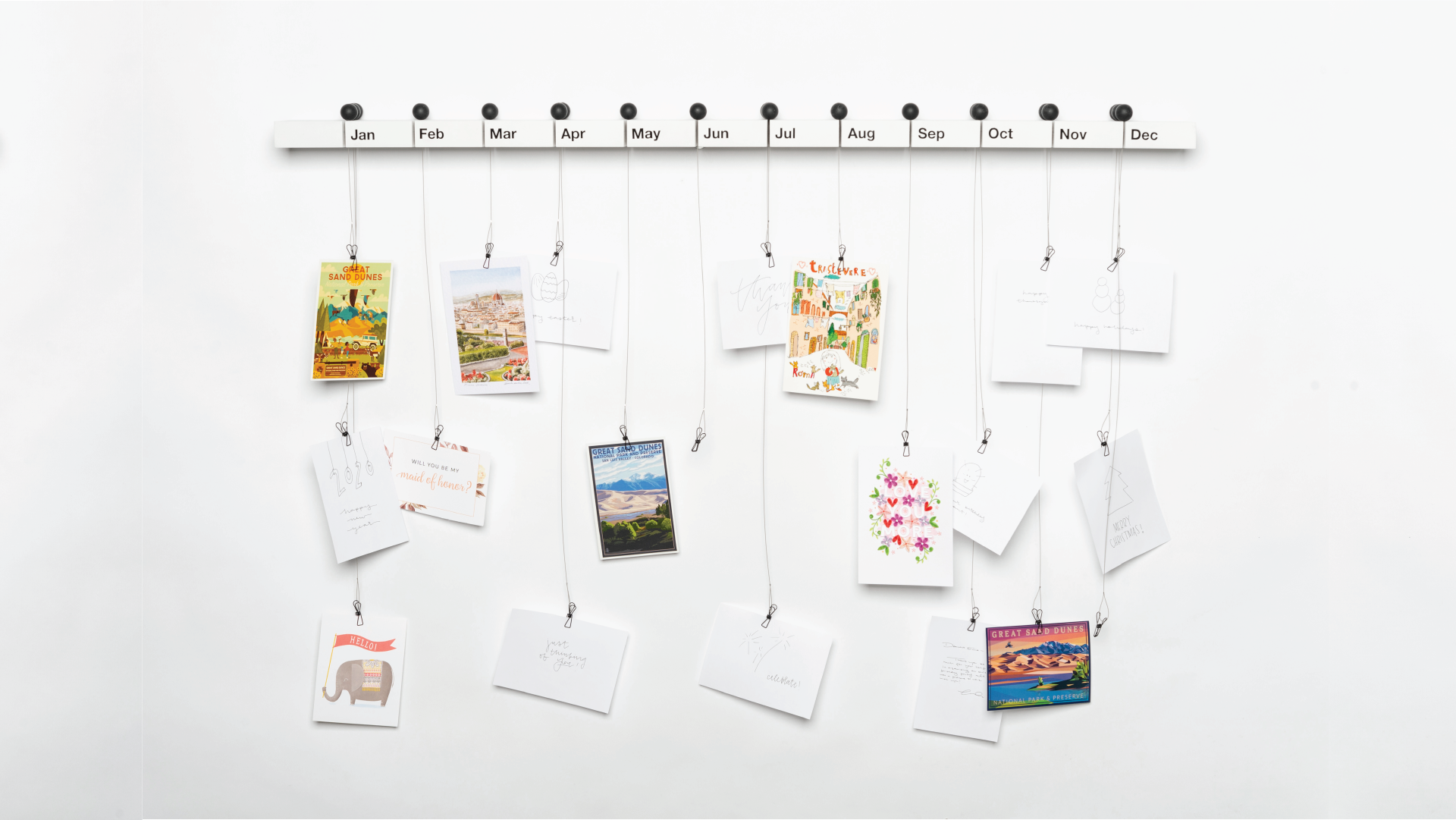 White shelf with vertical slots for each month of the year. Each slot has its own holder - a ball shape handle with a string attached to it. Photos and postcards are hanged through the black holders.