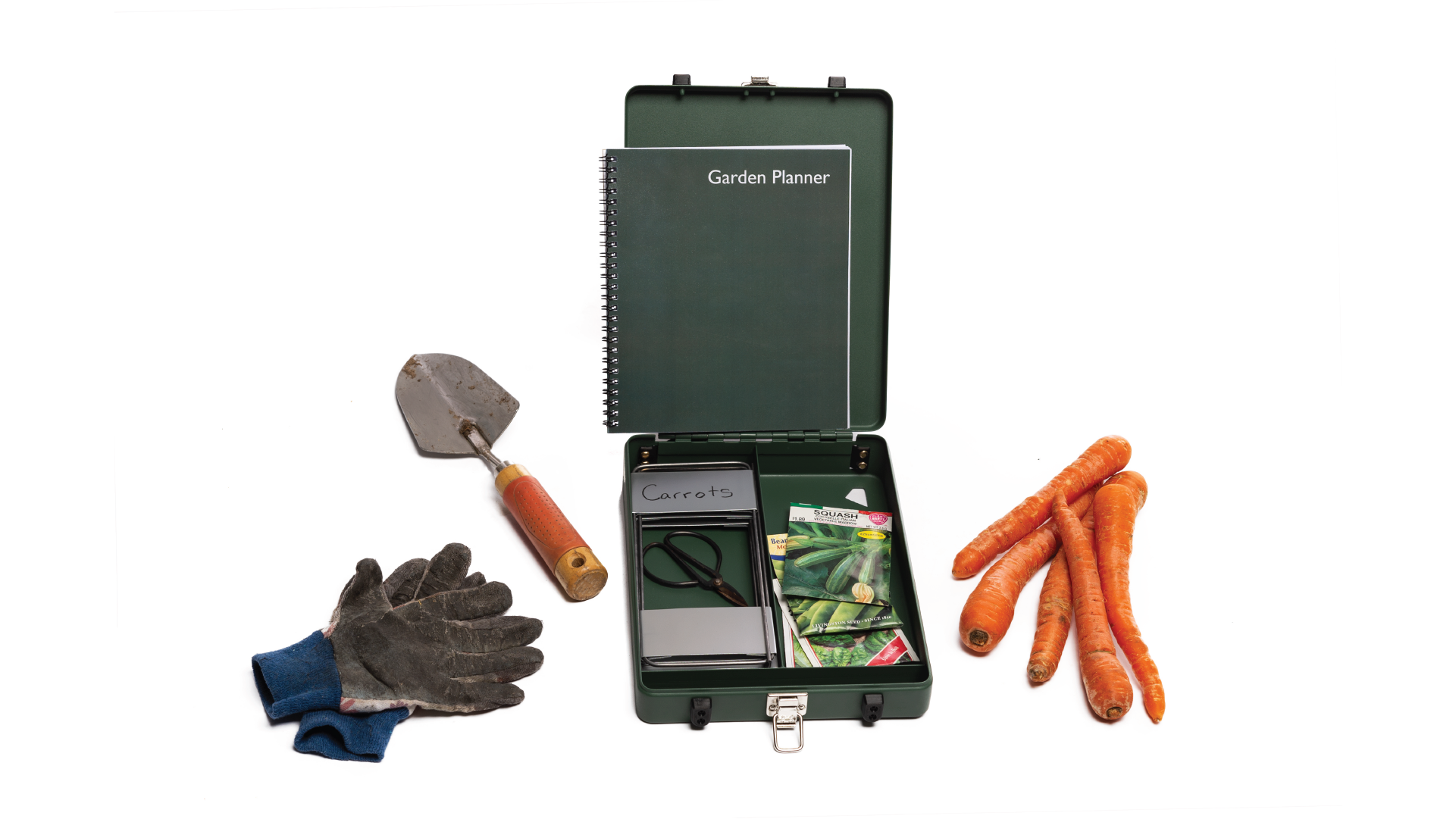 Green box open with gardening items inside. Outside the box there are gloves, a shovel and carrots (from left to right).