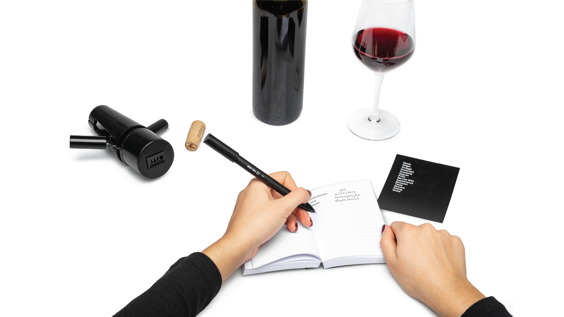 Hands writing on the accompanying journal with the corkscrew on the left, and a bottle and glass of wine on the right.