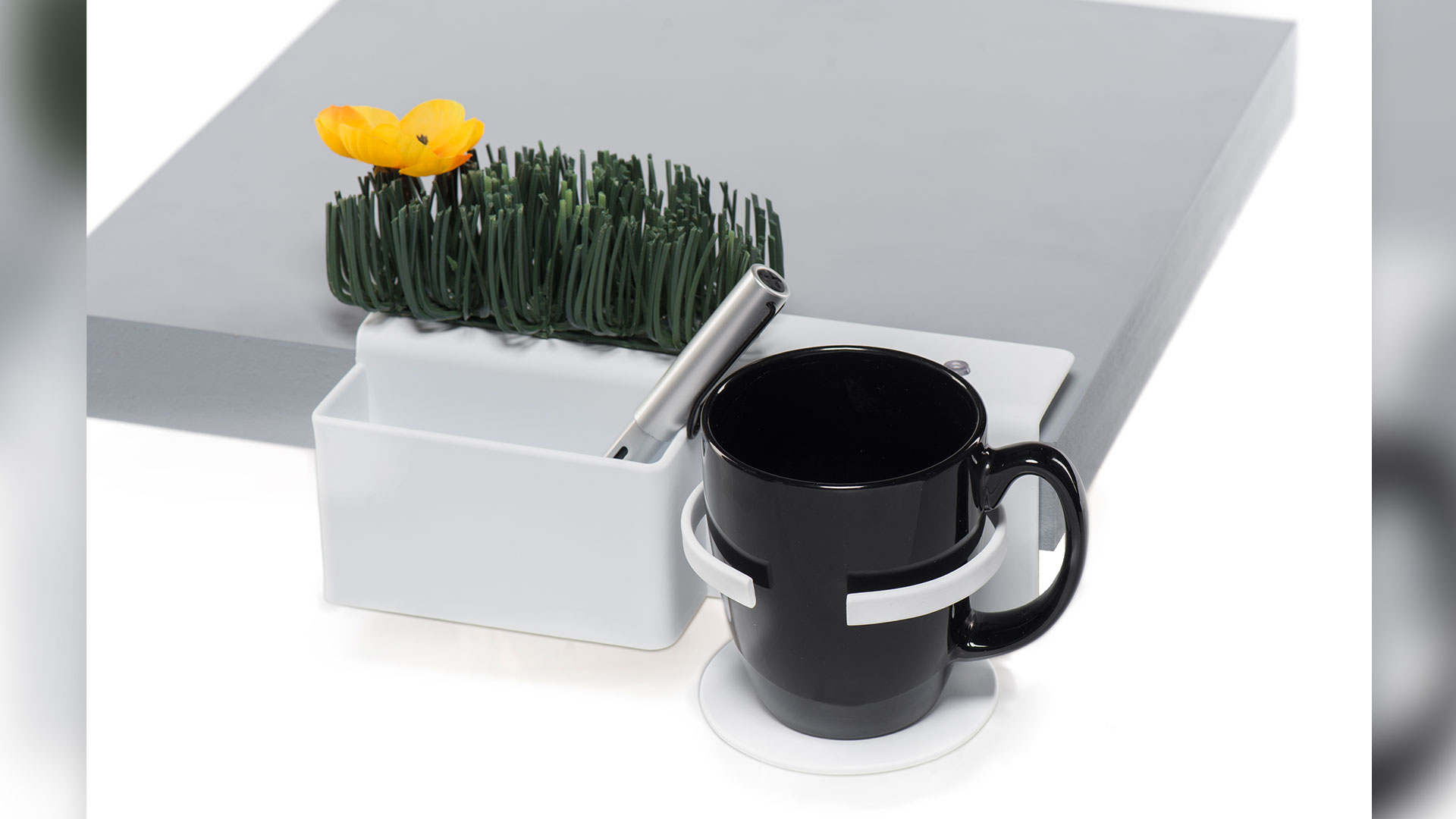 White Deskside shelf attached to side of table top a rectangular slot with pen inside, fake greenery above slot, a cup holder to the right of slot with black mug inside