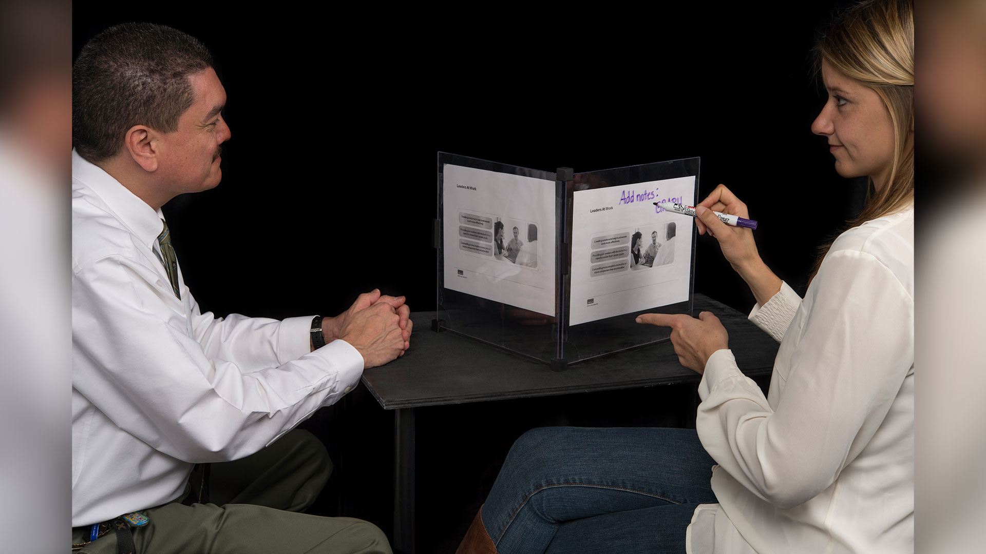 Two people seated at table looking at presentation cube, one with dry erase marker taking notes on top of presentation cube sheets