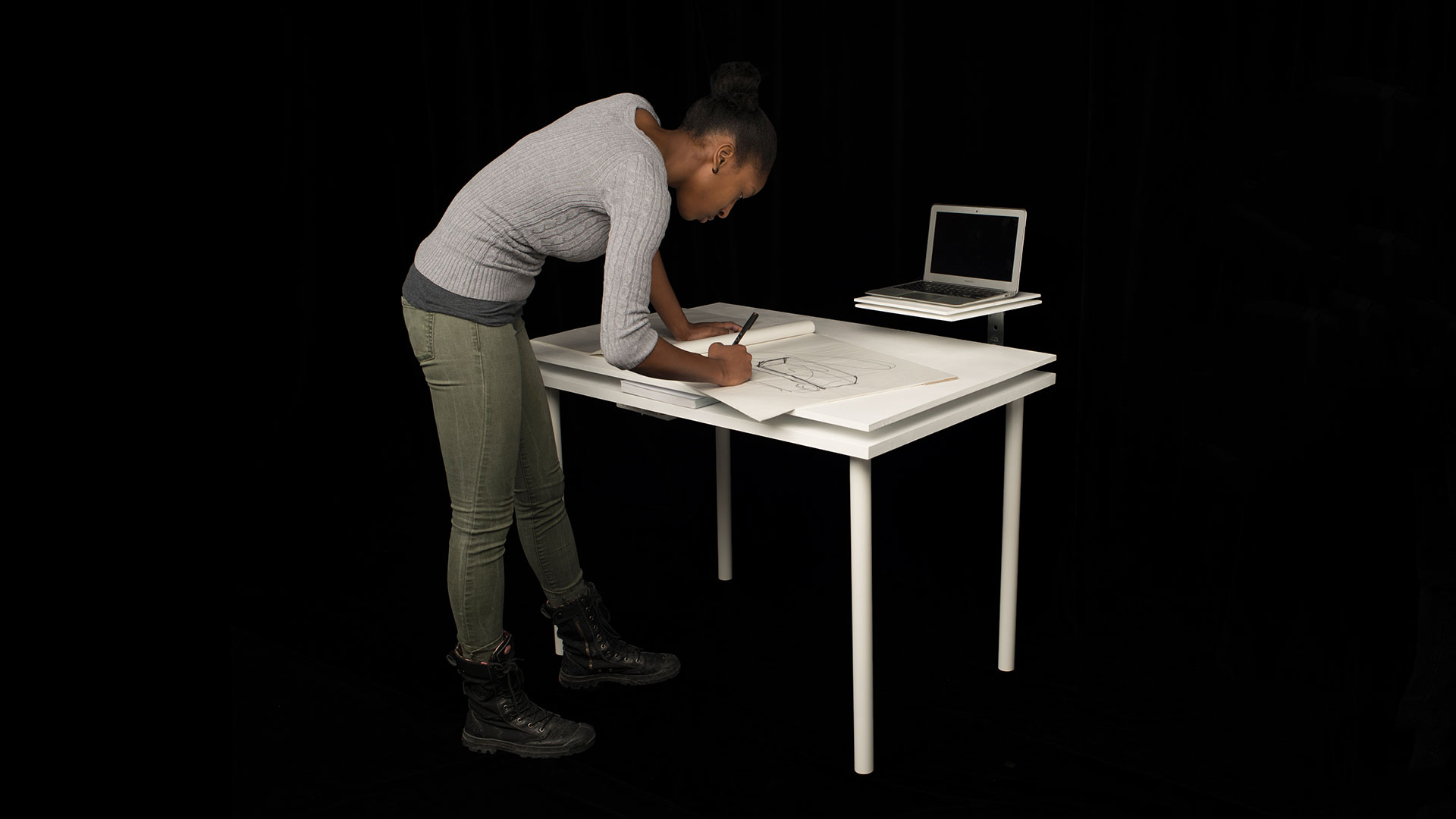 Student standing over table drawing on large drafting paper with pencil, a laptop sits on secondary shelf that attached via swivel component 