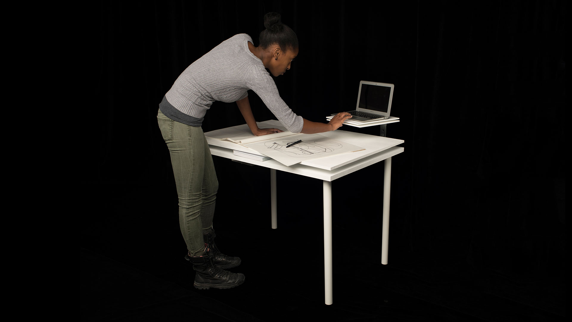 Student standing over table with drafting papers interacting with laptop that is on top of secondary swivel shelf