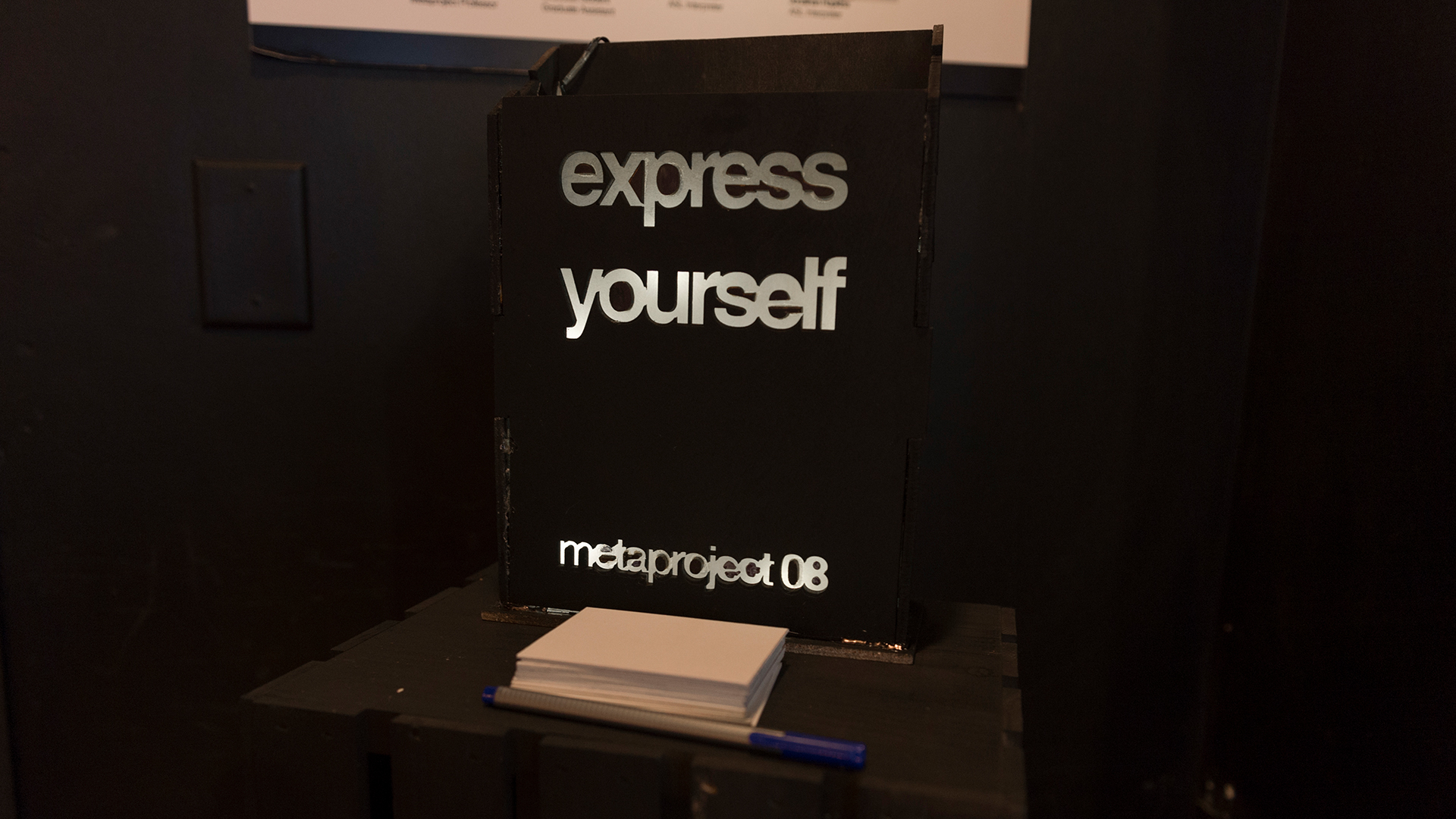 black sign with illuminated text on stand with stack of papers and pen
