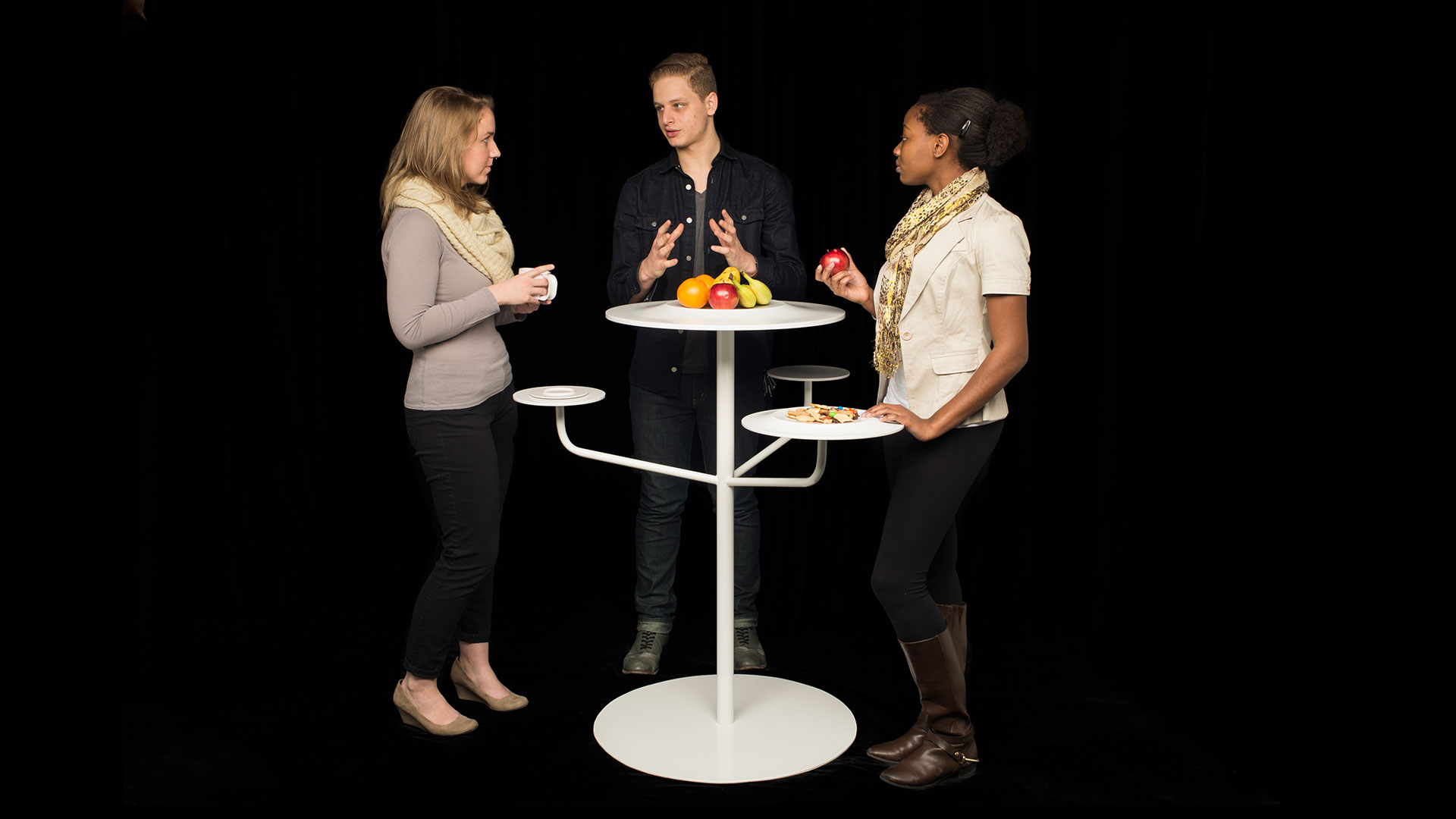 Three students gathered around a tall bar height circular white table with three smaller circular platforms coming out from central post below for additional surface area, assorted food items on table top