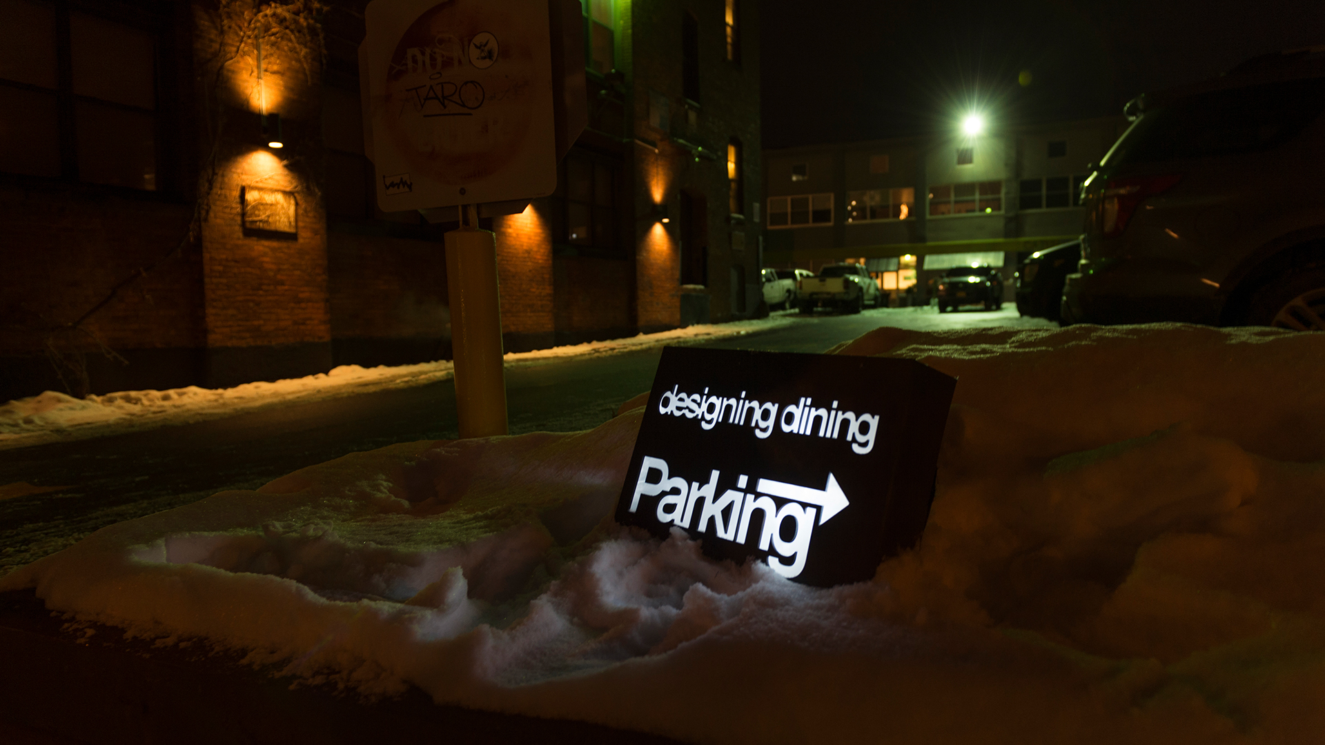 Exterior black sign with illuminated text sitting in snow pointing and with event name and designating parking location