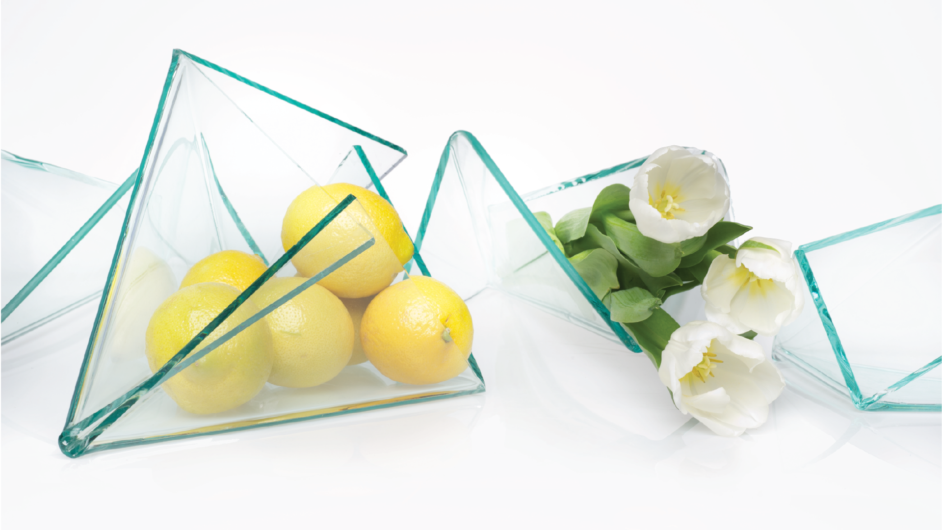 Closer shot of countertop two center-pieces made of folded glass sheets containing limes and flowers.