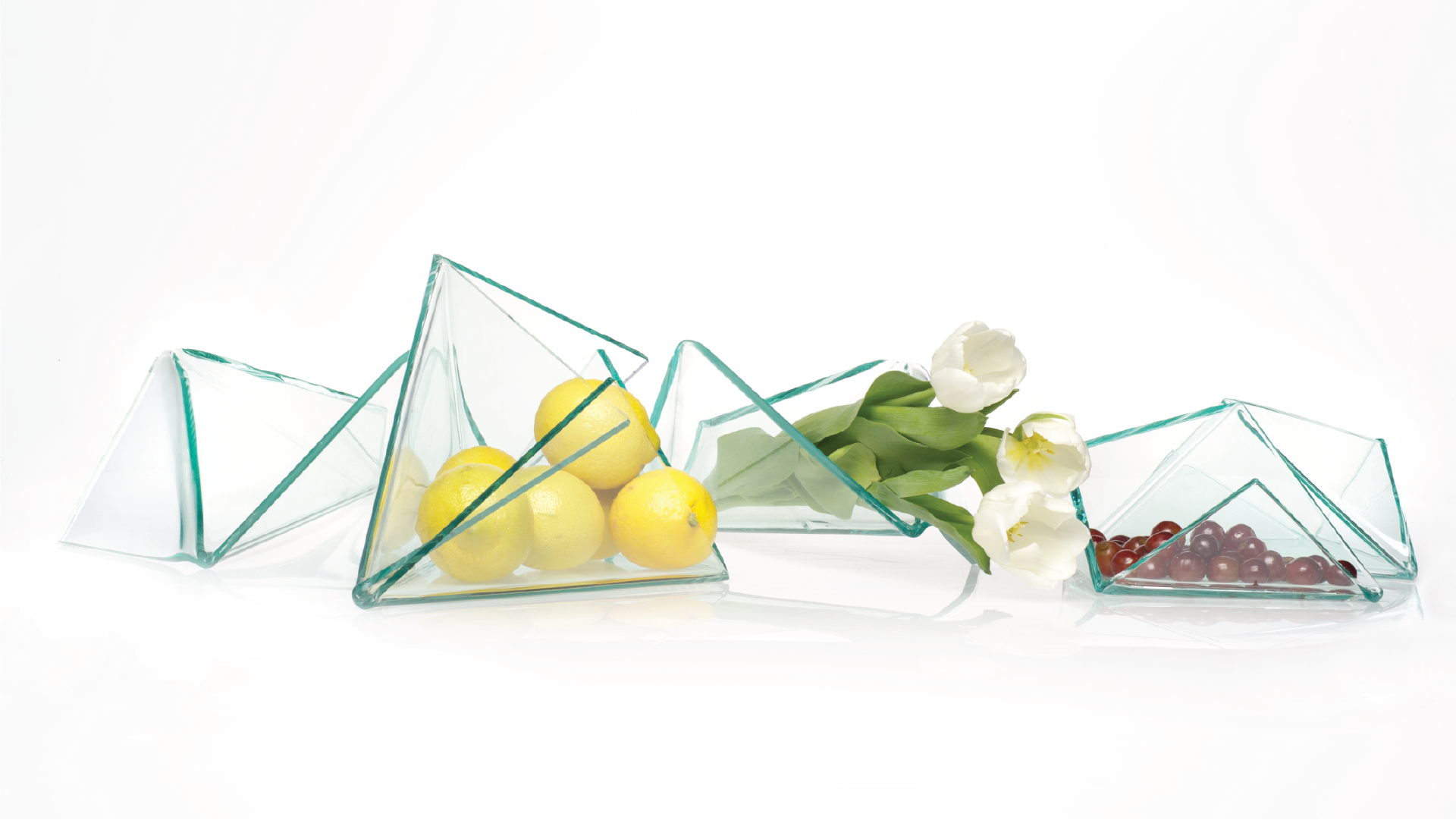 Four countertop center-pieces made of folded glass sheets containing limes, flowers and small fruits.