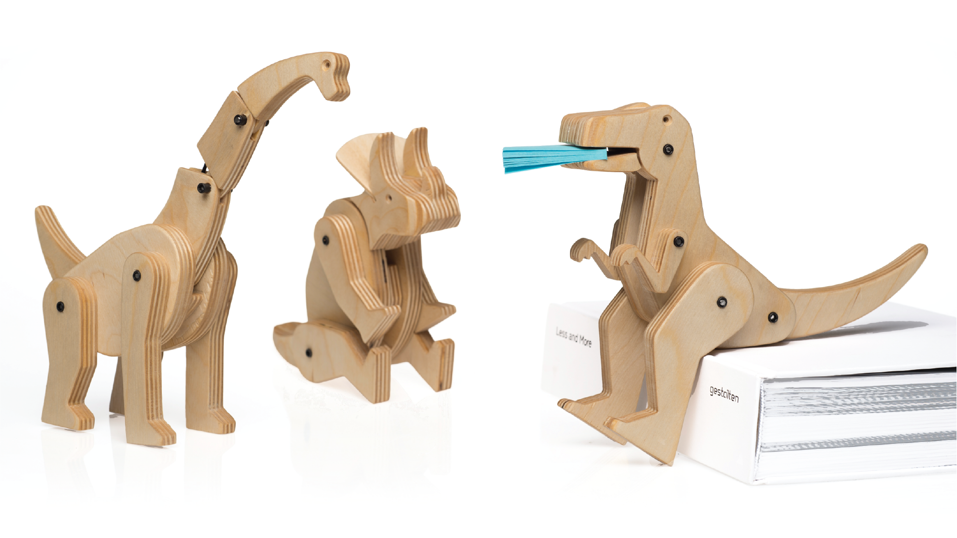3 wooden toys in different positions. One of the dinosaurs is sitting on a book and holds writing pad its mouth.