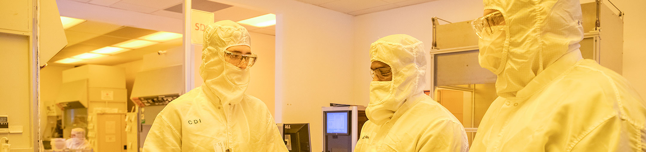 3 people in a clean room wearing lab suits