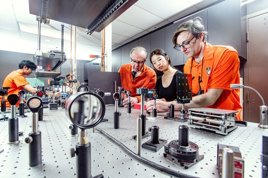A group of people in orange and black tops work in a lab.