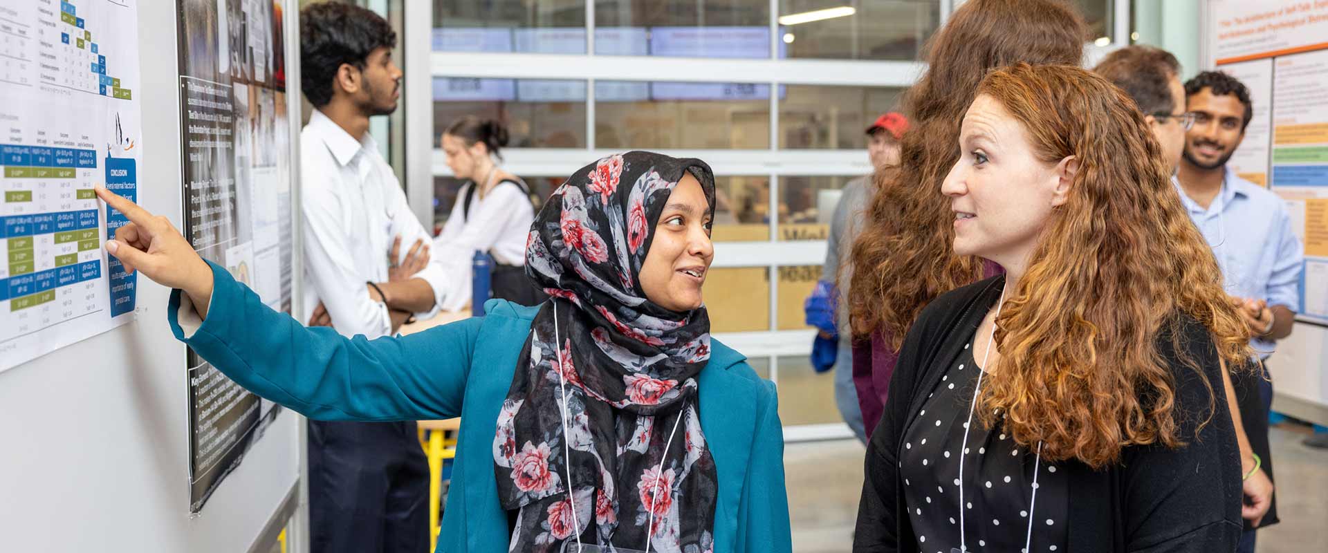 a student explains a poster presentation to a visitor at a research symposium.