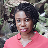 A woman in rose-colored blouse in stands in front of green foliage, smiling.