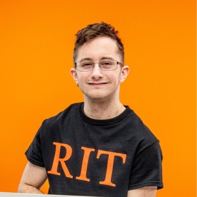 A man wearing glasses and a black shirt with orange letters that spell out RIT sits in front of an orange background, smiling.