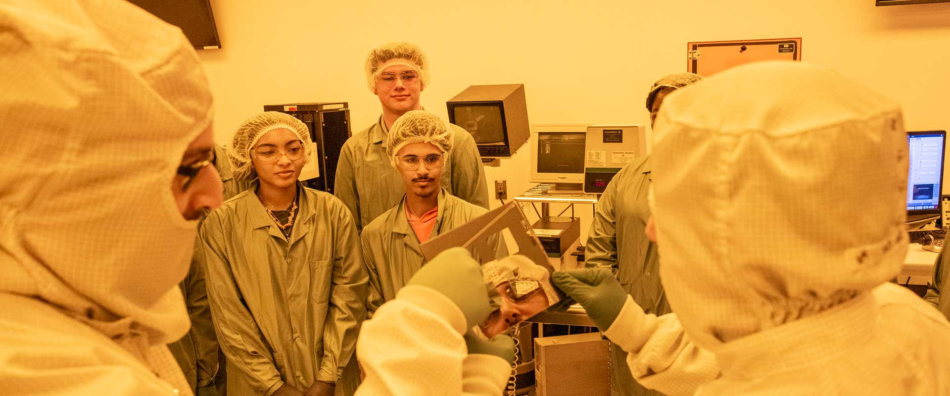 students and professors in a clean room, wearing protective gear and looking at semiconductors.