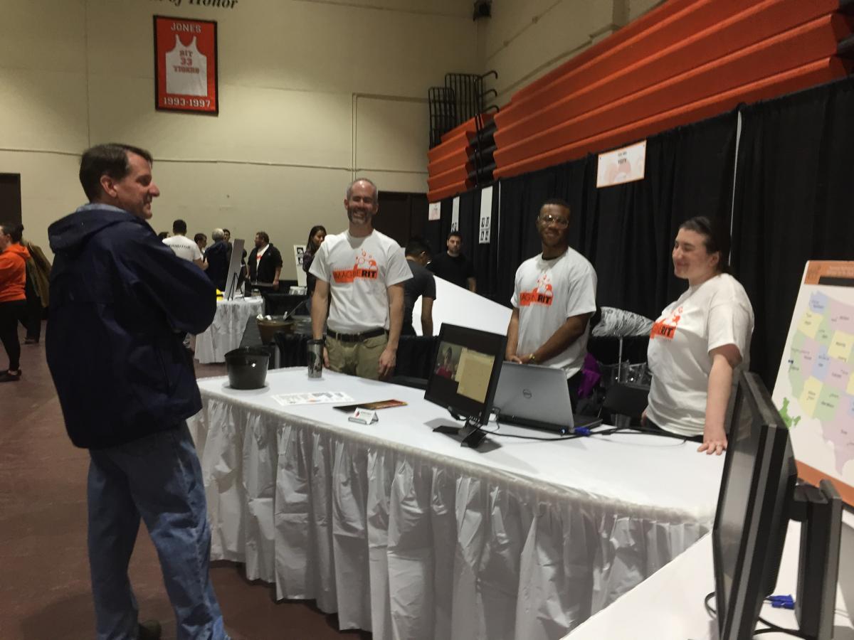 Three DHHVAC staff standing behind white table talking to Imagine RIT guest.