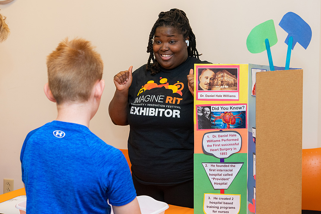 An exhibitor at ImagineRIT conversing with a young attendee