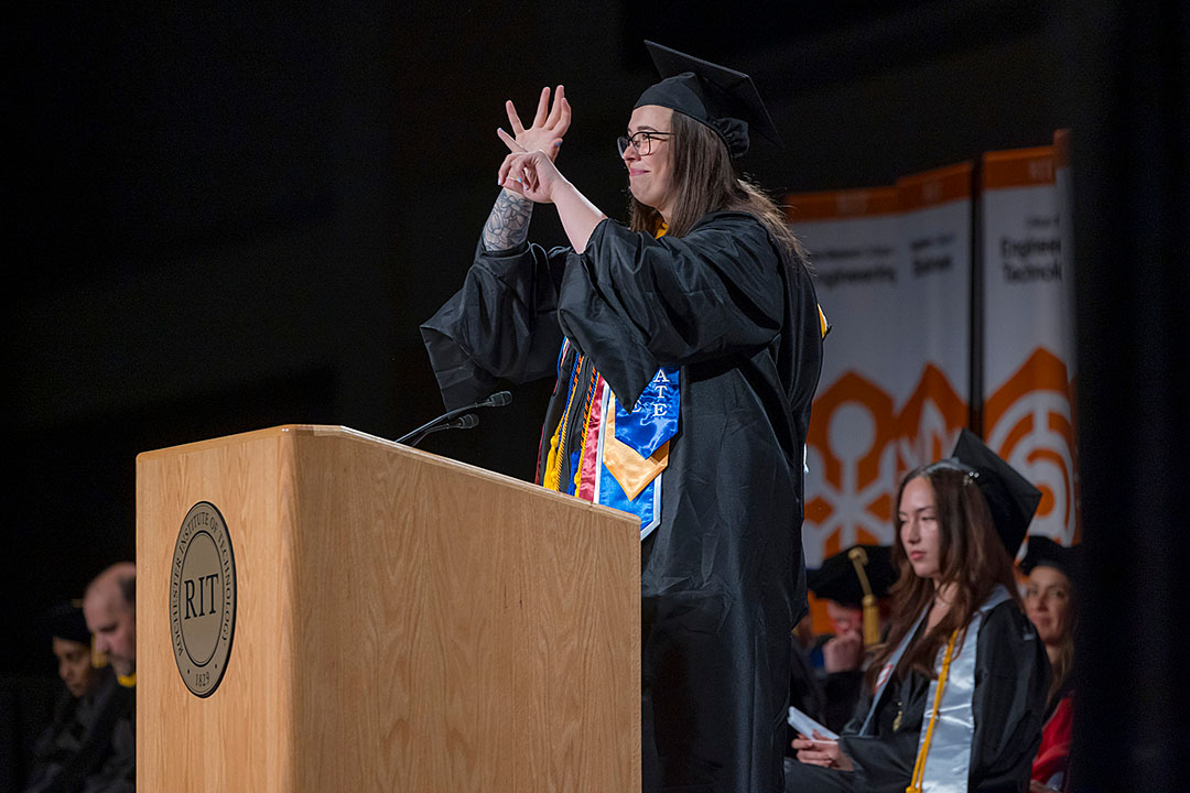 A graduate in a cap and gown stands at a podium with the RIT logo, using sign language during a commencement ceremony. Other graduates are seated in the background.