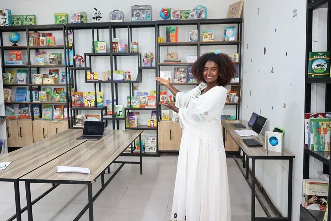 A smiling woman in a long white dress gestures toward bookshelves filled with books and learning materials in a modern study room.