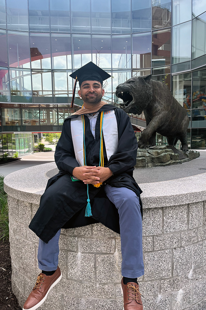 Dhruvesh Patel sitting on a stone wall in a graduation cap, gown, and academic stoles in front of the bronze RIT tiger statue.
